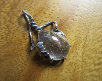 Puede incluir: Pendiente de plata con una piedra de cuarzo transparente con inclusiones doradas, engastada en un diseño de alambre de plata trenzado.