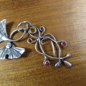 Puede incluir: Un colgante de plata con un dise&ntilde;o estilizado de hoja de ginkgo con una hoja m&aacute;s peque&ntilde;a y tres piedras preciosas. Las piedras preciosas son naranja, roja y roja.