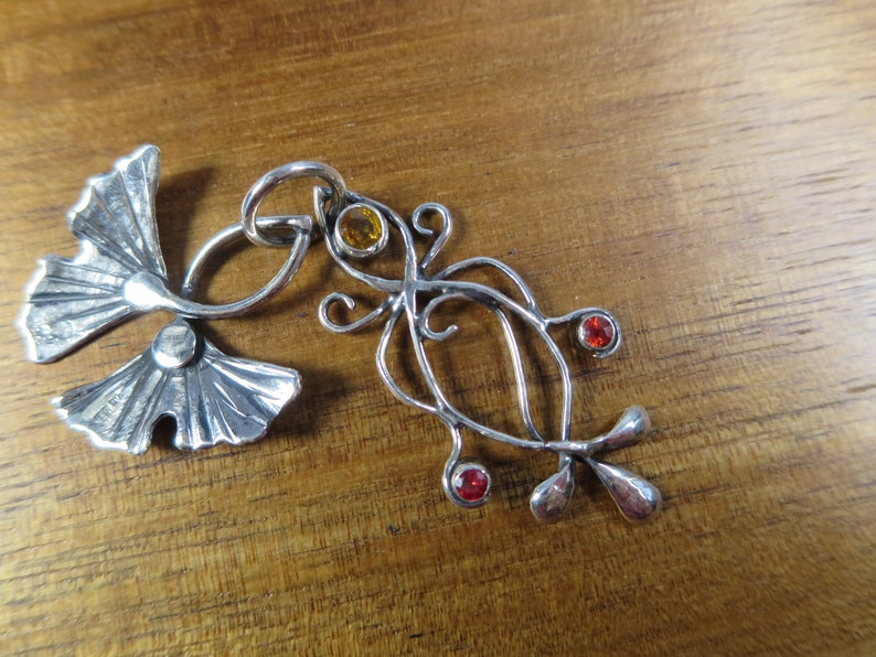 Puede incluir: Un colgante de plata con un dise&ntilde;o estilizado de hoja de ginkgo con una hoja m&aacute;s peque&ntilde;a y tres piedras preciosas. Las piedras preciosas son naranja, roja y roja.