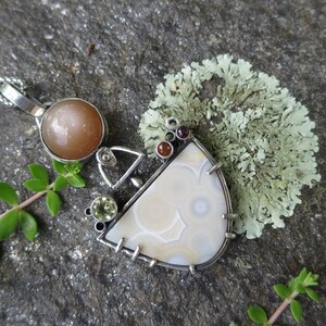 May include: A silver pendant necklace with a white agate stone, a sunstone, and various other gemstones. The pendant is set in a silver frame and hangs from a silver chain.