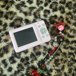 May include: A pink digital camera with a black screen and silver buttons. The camera is lying on a brown and white leopard print fabric. A beaded keychain with red, black, and silver beads, a cherry charm, and a silver charm is hanging from the camera.