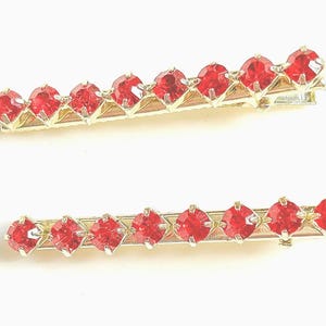 Può includere: Due fermagli per capelli dorati con dettagli in strass rossi. Ogni fermaglio ha una fila di strass rossi.