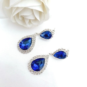 Könnte beinhalten: Ein Paar Tropfenohrringe mit großen, tropfenförmigen, tiefblauen Edelsteinen. Jeder Ohrring ist von kleinen, klaren Kristallen umgeben, die in silberfarbenem Metall gefasst sind. Eine weiße Rose ist im Hintergrund.