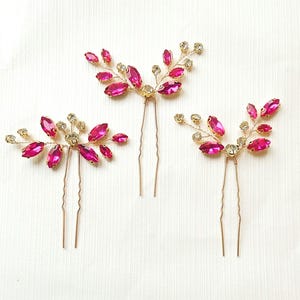 May include: Three decorative hair pins with a floral design. Each pin features a gold-coloured wire base adorned with bright pink, oval-shaped crystals and smaller clear crystals. The pins are arranged on a white surface.