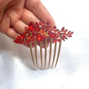 May include: A gold-coloured hair comb decorated with bright red gemstones. The comb has a floral design with red crystals of different sizes and shapes, creating a sparkling accessory. The comb's teeth are evenly spaced.