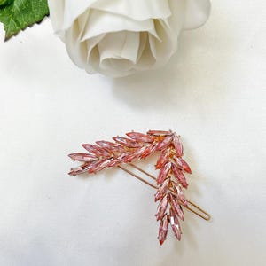 May include: Two pink crystal hairpins in a wheat stalk design. The hair accessories are made of small, pink crystals set on a gold-toned metal base. A white rose is in the background.