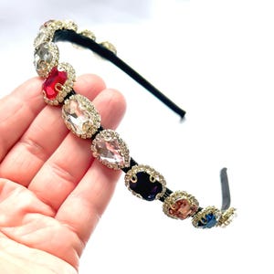 May include: A black headband featuring a line of multi-coloured, teardrop-shaped rhinestones. The rhinestones, in shades of red, clear, pink, and blue, are set in gold-coloured metal.