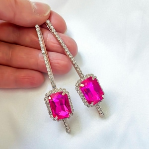 May include: Two hairpins featuring a rectangular, magenta gemstone centre, encircled by a halo of small, clear crystals. The pins are long and slender, with a line of small crystals along their length.