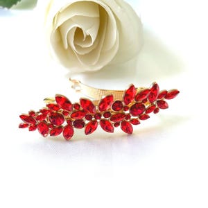 Può includere: Un fermaglio per capelli dorato ornato da numerosi abbellimenti rossi a forma di gioiello. Il fermaglio presenta un design a forma di foglia con pietre rosse di varie dimensioni. Una rosa bianca è sullo sfondo.