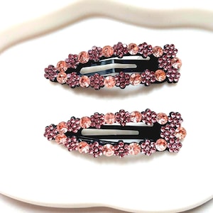 May include: Two black hair clips with pink and light pink rhinestones arranged in a flower pattern.