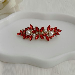 May include: A red and pearl-accented hair clip. The clip features red, leaf-shaped crystals and several white pearls. The clip is set on a gold-toned metal base. The hair accessory is designed for formal occasions.