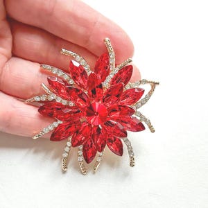 Puede incluir: Broche de metal dorado con forma de flor, con grandes pétalos de cristal rojo y un cristal rojo central. El broche está adornado con cristales transparentes y tiene un cierre de alfiler. Un accesorio decorativo.