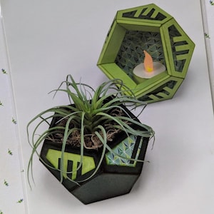 May include: A Sizzix Thinlits die set by Lynda Kanase, featuring geometric shapes for crafting. The image shows a finished papercraft project: a black and green geometric planter with a small plant and a matching candle holder.