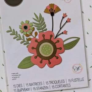 May include: A package of Sizzix Thinlits dies featuring a floral design. The die-cut flowers are in shades of coral, green, and brown. The package includes the text "Sizzix", "Thinlits", and "Olivia Rose". The package also states "15 Dies".