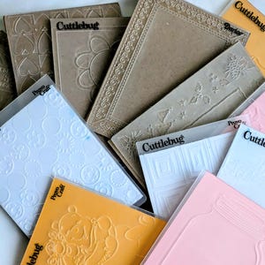 May include: A collection of Cuttlebug embossing folders in various colors and designs. The folders feature embossed patterns including flowers, geometric shapes, and Winnie the Pooh. Some folders are tan, white, pink, and orange.