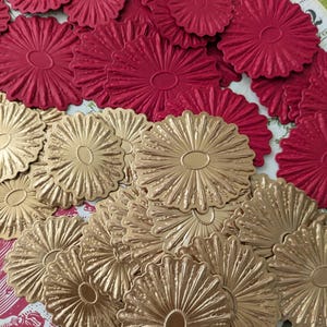 May include: A collection of embossed paper die cuts in a flower shape. The die cuts are in two colors: a deep red and a metallic gold. The flower shapes have a central oval and radiating lines.