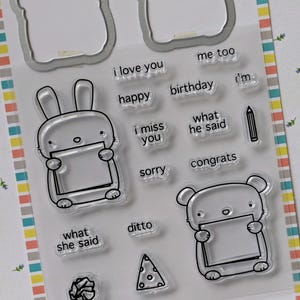 May include: Clear stamp set with cartoon bunny and bear characters holding blank signs. Includes stamps with phrases like "I love you," "Happy Birthday," and "Congrats." Also features a pencil and other small images.