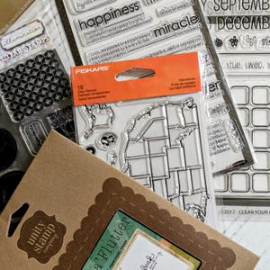 May include: A collection of clear stamps for crafting, scrapbooking, and journaling. Includes stamps with phrases like "happiness" and "miracle", a map of the United States, and calendar elements. Packaging includes the text "Books a'Flutter".