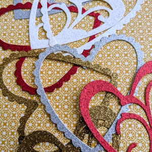 May include: An assortment of glittery heart-shaped cutouts in red, silver, and gold, scattered on a yellow patterned background. The hearts have decorative edges and intricate openwork designs, perfect for crafting or Valentine's Day decorations.