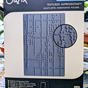 May include: A Sizzix Seth Apter Textured Impressions multi-level embossing folder in its original packaging. The gray plastic folder features a grid-like pattern with circular and linear raised details, shown in a circular inset preview.