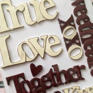 May include: Close-up of decorative wooden words and phrases. The words "True Love" and "Hugs & Kiss" are in a cream color, while "Together" and "XOXO" are in a dark red. A small red heart is also present.