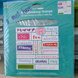 May include: A teal package of Night Media Scrapbooking Stamps. The package contains 22 clear stamps for perfect positioning. The stamps feature words like "funny", "chic", "strong", and "adorable". The text "a must have" is written on the package.