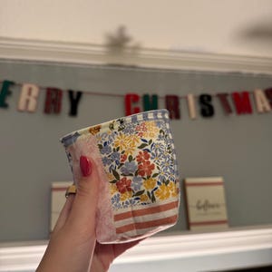 May include: A small, cylindrical fabric basket featuring a patchwork design with various floral patterns in yellow, red, and blue, accented by sections of pink fabric and horizontal stripes, finished with a light blue and white gingham trim.
