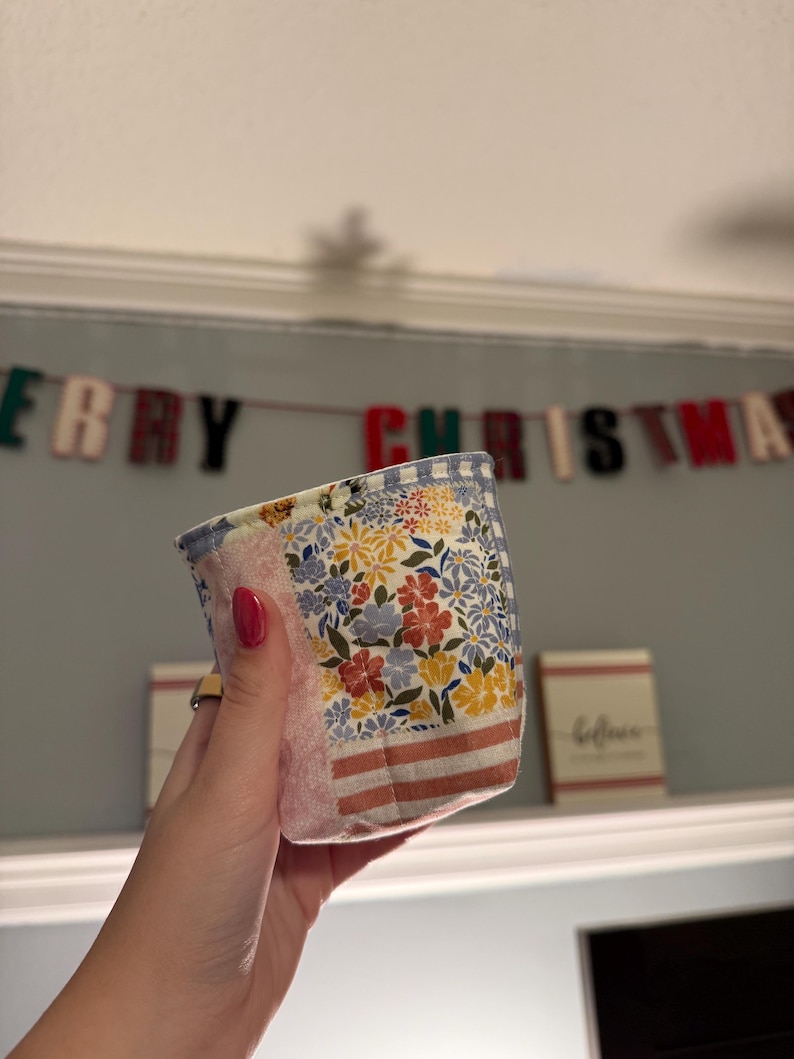 May include: A small, cylindrical fabric basket featuring a patchwork design with various floral patterns in yellow, red, and blue, accented by sections of pink fabric and horizontal stripes, finished with a light blue and white gingham trim.