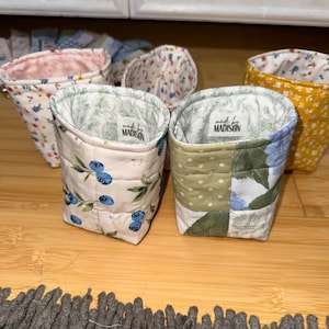 May include: A collection of four small, quilted fabric pouches with open tops. Each pouch features different floral or fruit patterns, including blueberries and green leaves. Two pouches have a white label inside reading made by MADISON.