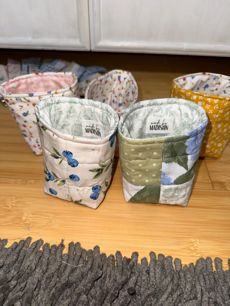 May include: A collection of four small, quilted fabric pouches with open tops. Each pouch features different floral or fruit patterns, including blueberries and green leaves. Two pouches have a white label inside reading made by MADISON.