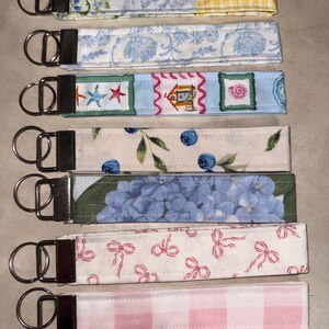 May include: Seven fabric wristlet keychains with silver-tone metal hardware and split rings. The patterns include yellow gingham, blue floral, beach motifs, blueberries, blue hydrangeas, pink bows, and a pink and white gingham check design.