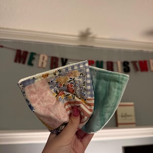 May include: A fabric bowl cozy featuring a patchwork design with floral, striped, and gingham patterns in shades of pink, blue, and yellow. The reverse side is a solid light green fabric with a quilted texture and visible stitching throughout.