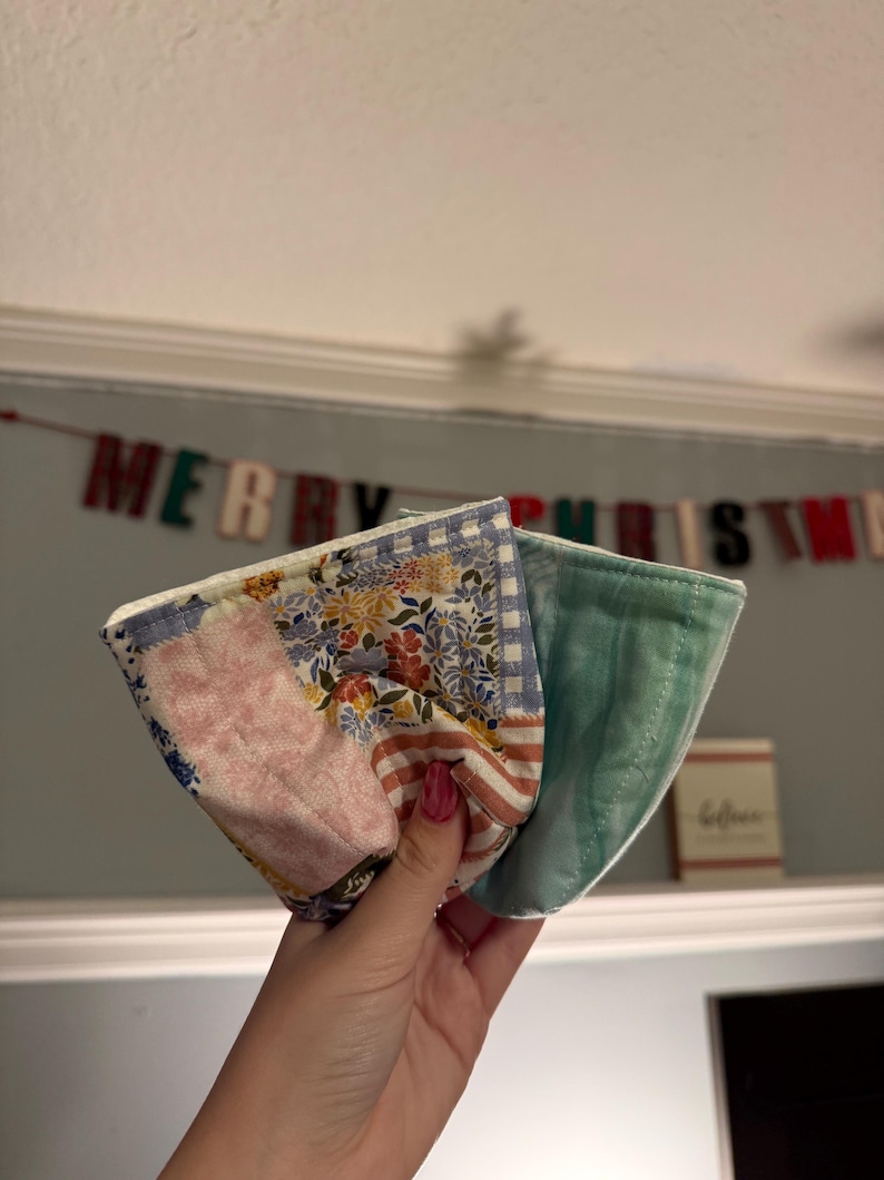 May include: A fabric bowl cozy featuring a patchwork design with floral, striped, and gingham patterns in shades of pink, blue, and yellow. The reverse side is a solid light green fabric with a quilted texture and visible stitching throughout.