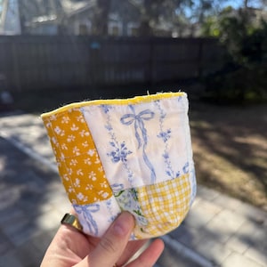 May include: A quilted fabric cup cozy featuring a patchwork design of mustard yellow floral print, white fabric with blue ribbon and floral motifs, and a yellow and white gingham check pattern, finished with a bright yellow fabric trim edge.