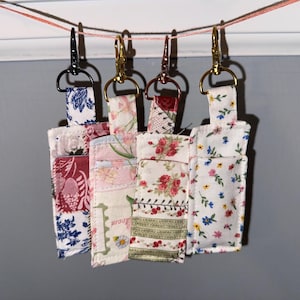 May include: Four rectangular fabric pouches with floral patterns hanging from a string by metal lobster clasps. The pouches feature various prints including blue and white toile, pink floral, red rose, and small multi-colored flower designs.