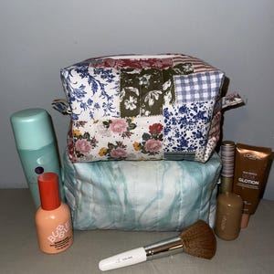 May include: A patchwork cosmetic bag with floral and patterned fabric sits atop a light blue quilted bag. Makeup products, including a brush, spray bottle, and tubes of lotion, are arranged around the bags.