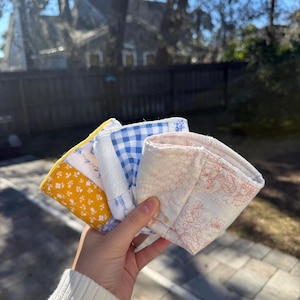May include: A set of three fabric cup sleeves featuring different patterns: one with a yellow and white floral print, one with a blue and white gingham check, and one with a light pink and white floral line drawing on a cream background.