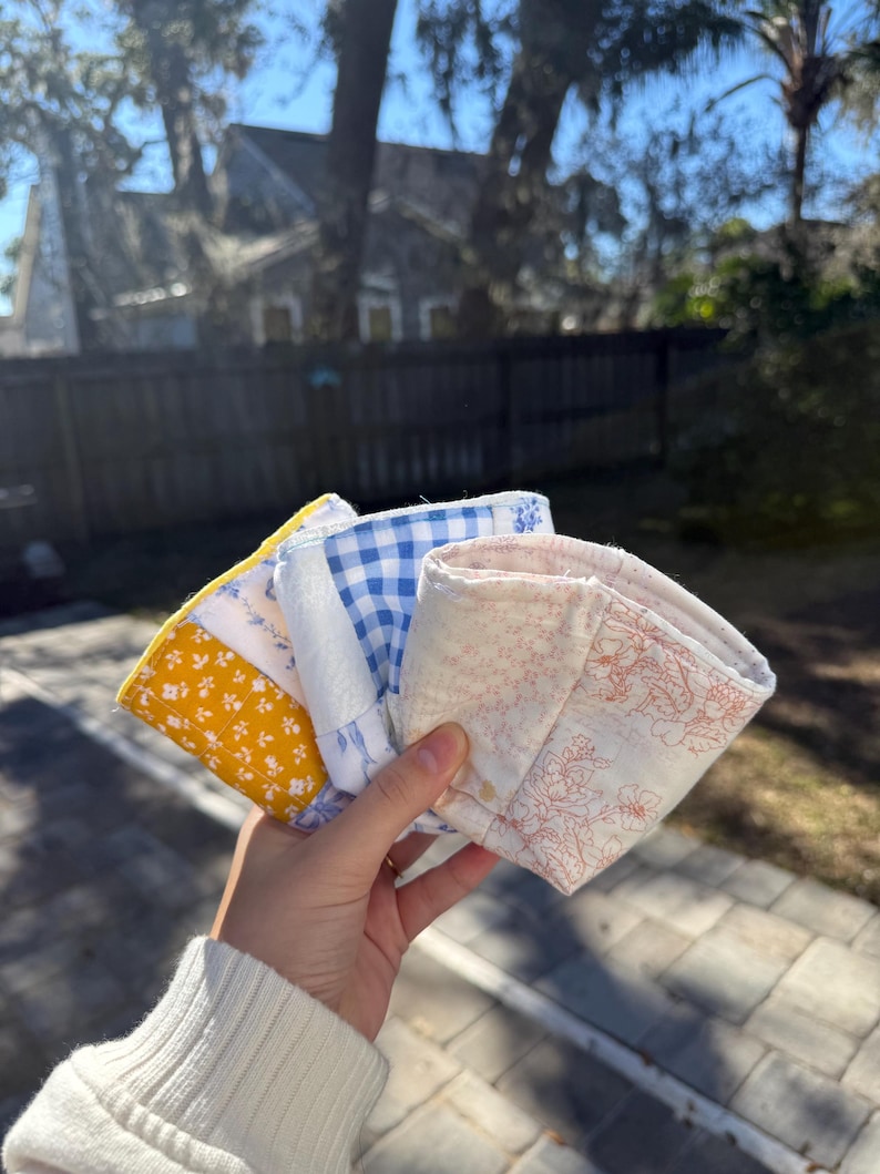 May include: A set of three fabric cup sleeves featuring different patterns: one with a yellow and white floral print, one with a blue and white gingham check, and one with a light pink and white floral line drawing on a cream background.