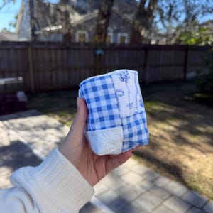 May include: A small, cylindrical fabric container featuring a patchwork design of blue and white gingham check patterns, a white fabric with a blue floral print, and a textured white fabric panel, stitched together in a quilted construction.