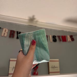May include: A small, rectangular fabric pouch featuring a marbled pattern in shades of light green, teal, and white. The fabric has visible vertical stitching lines and a soft, folded construction, presenting a smooth texture throughout the surface.