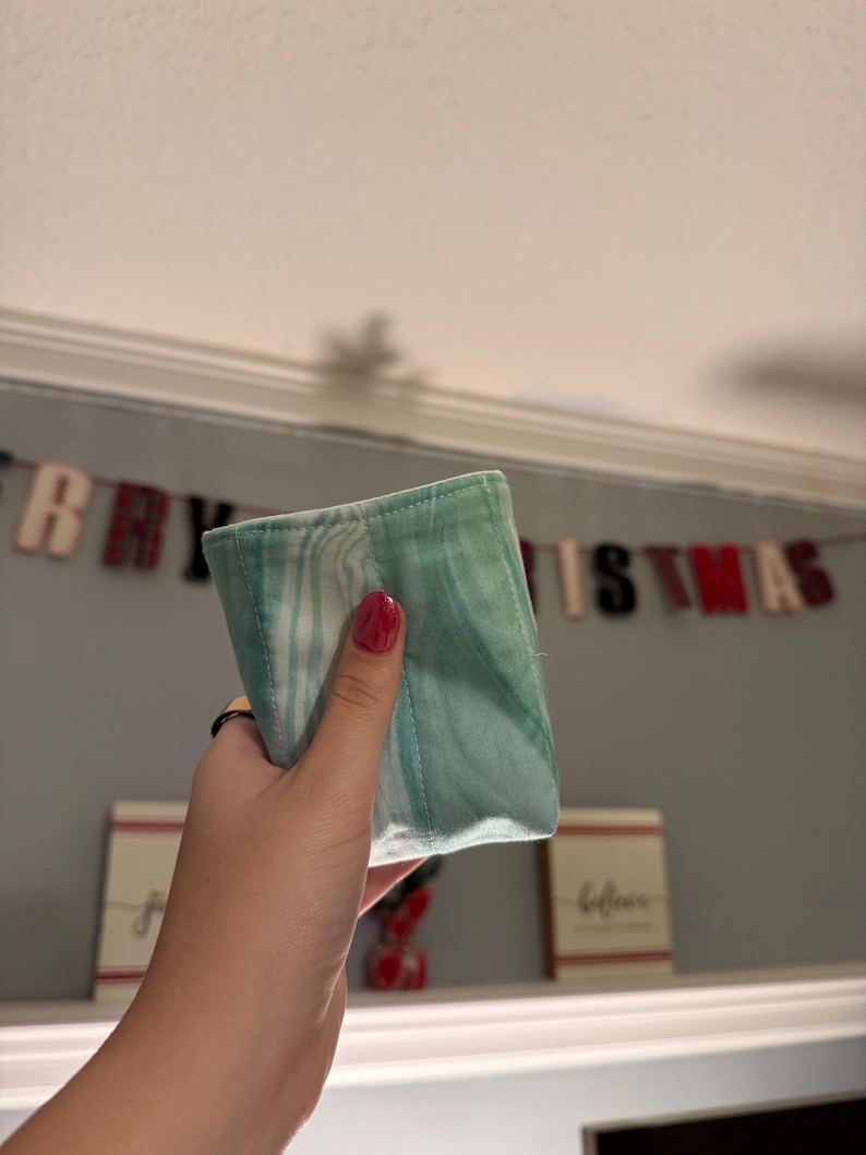 May include: A small, rectangular fabric pouch featuring a marbled pattern in shades of light green, teal, and white. The fabric has visible vertical stitching lines and a soft, folded construction, presenting a smooth texture throughout the surface.