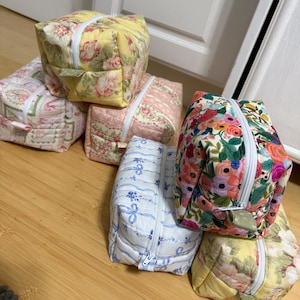 May include: A collection of five rectangular quilted fabric pouches featuring various floral and ribbon patterns in yellow, pink, blue, and multi-colored designs. Each pouch is constructed with a white zipper closure and a small fabric pull tab.