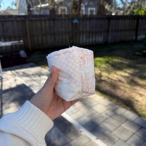May include: A small, quilted fabric pouch featuring a patchwork design with a light cream background. One side displays a delicate pink floral pattern, while the adjacent side showcases a detailed orange-toned botanical line art illustration.