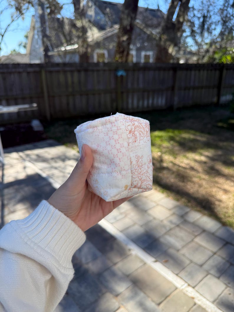 May include: A small, quilted fabric pouch featuring a patchwork design with a light cream background. One side displays a delicate pink floral pattern, while the adjacent side showcases a detailed orange-toned botanical line art illustration.