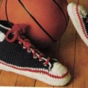 Puede incluir: Zapatillas altas de ganchillo en azul marino, blanco y rojo. Las zapatillas tienen punteras blancas, cordones rojos y un círculo blanco en el lateral. Un balón de baloncesto está en el fondo.