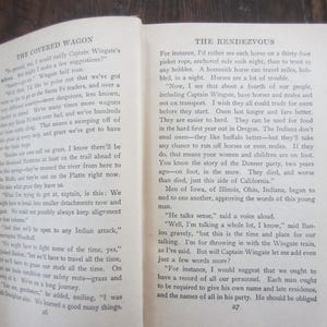 Puede incluir: Primer plano de dos p&aacute;ginas abiertas de un libro titulado "The Covered Wagon" y "The Rendezvous". El texto trata sobre los desaf&iacute;os de viajar en caravana de carros, incluyendo la necesidad de campamentos seguros y la importancia de tener bueyes en lugar de caballos.
