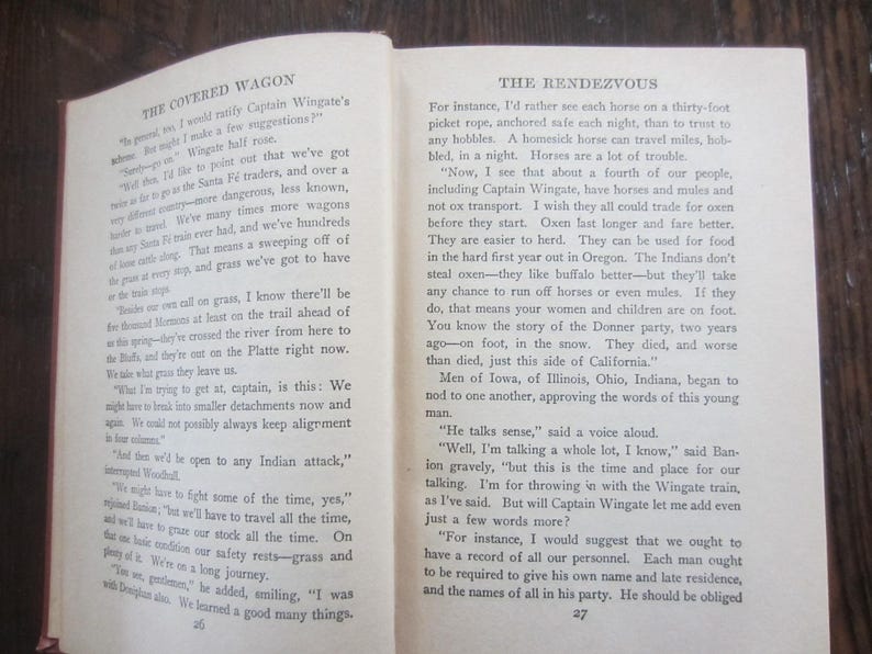 Puede incluir: Primer plano de dos p&aacute;ginas abiertas de un libro titulado "The Covered Wagon" y "The Rendezvous". El texto trata sobre los desaf&iacute;os de viajar en caravana de carros, incluyendo la necesidad de campamentos seguros y la importancia de tener bueyes en lugar de caballos.