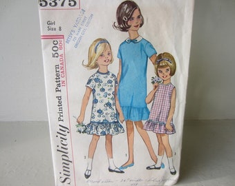 May include: Simplicity Printed Pattern 500 in Canada 60c. A vintage sewing pattern for a girl's dress. The pattern shows three girls wearing different styles of dresses. The dress on the left is a white dress with blue floral print. The dress in the middle is a light blue dress. The dress on the right is a pink and white gingham dress.