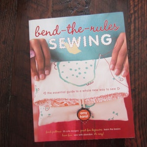 May include: A book titled "bend-the-rules SEWING" with a colorful fabric project on the cover. The book's subtitle is "the essential guide to a whole new way to sew". The author's name, "amy karol", is printed on a red circle on the cover. The book's description reads "fresh patterns 30 cute designs great for beginners learn the basics have fun sew with abandon it's easy"