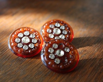 May include: Three round, brown buttons with a raised design of clear rhinestones.